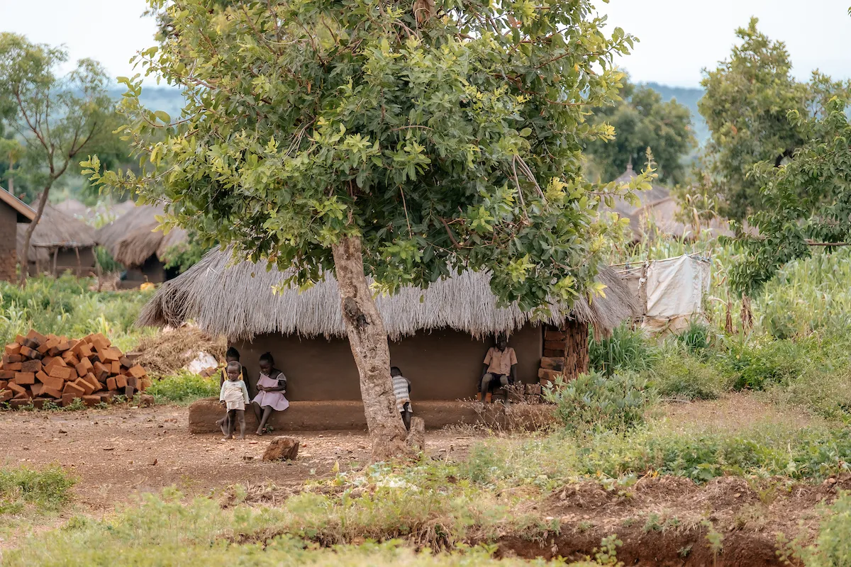 Ugandan refugee children sitting in front of a thatched-roof home.