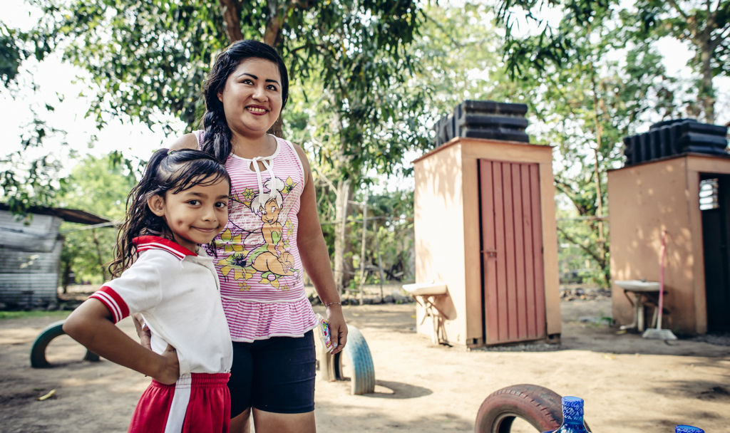 Ximena, 6-years-old, stands next to her mother outside of their pit latrines, which used to seep into their water source. El Encanto, Mexico